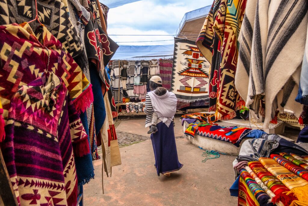 Un siglo de intercambio: El pulso vivo de la Feria Dominical de Huancayo Colorful display of traditional textiles at a bustling outdoor market with a local shopper.
