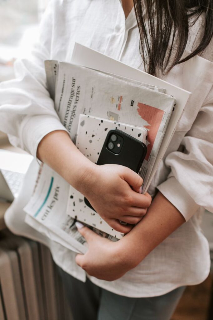 A woman in a white shirt holds newspapers and a smartphone indoors, standing by a window.